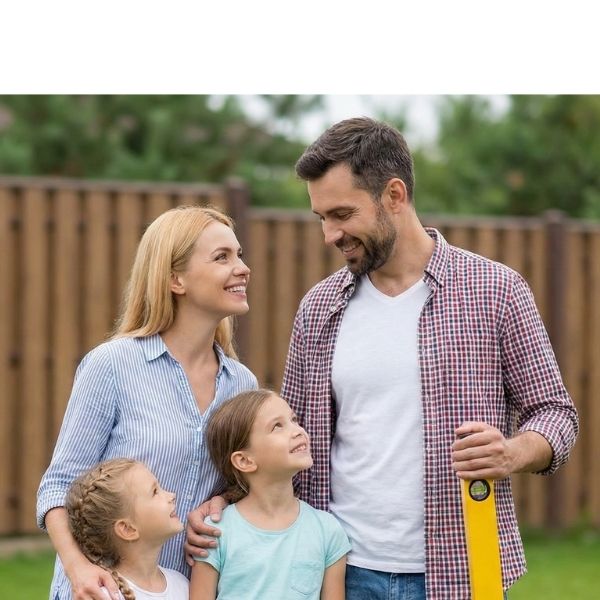 Eine vierköpfige Familie (Vater, Mutter und zwei Töchter) steht lächelnd im Freien vor einem Holzlattenzaun. 