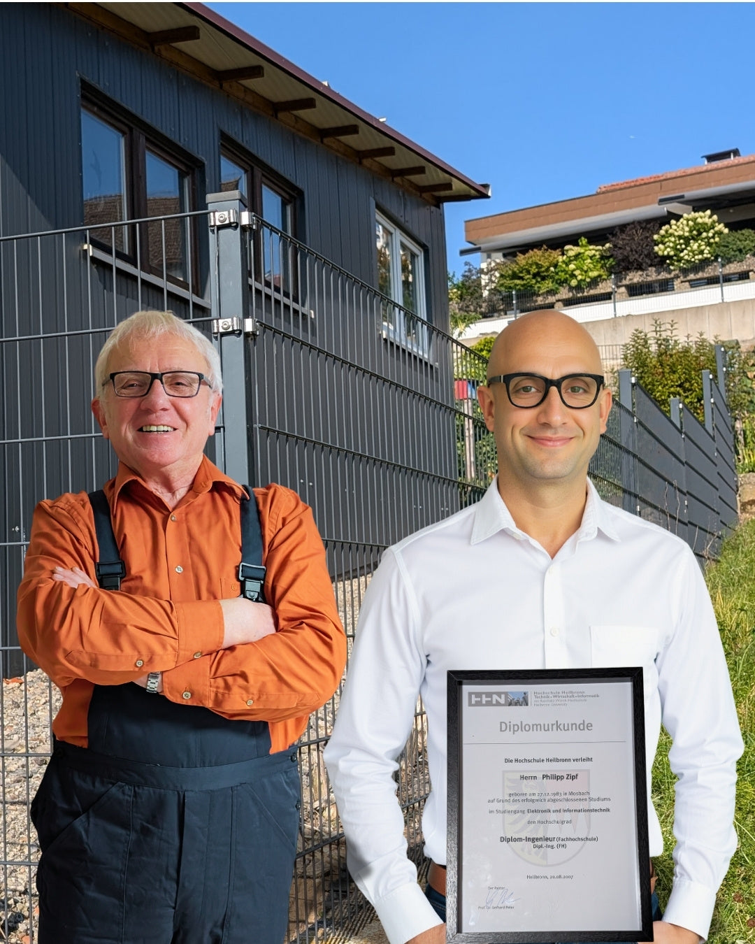 Ein Teamfoto von Vater und Sohn vor einem modernen, dunklen Doppelstabmattenzaun von Qualinos.